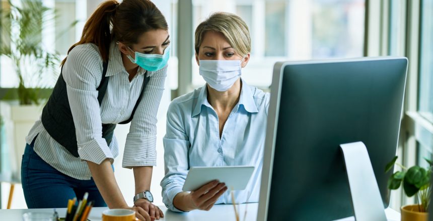 Female entrepreneurs with face masks cooperating while working on touchpad in the office.